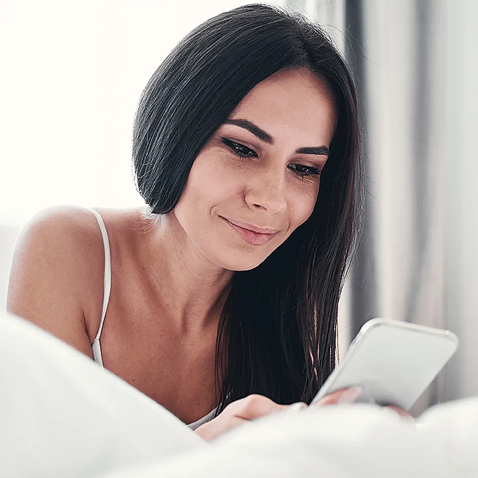 Fiatal nő mobiltelefonon keresztül időpontot foglal lézeres végleges szőrtelenítésre, otthoni környezetben, mosolyogva